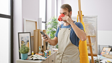 A bearded man in an apron enjoys coffee while checking his phone in a bright art studio.
