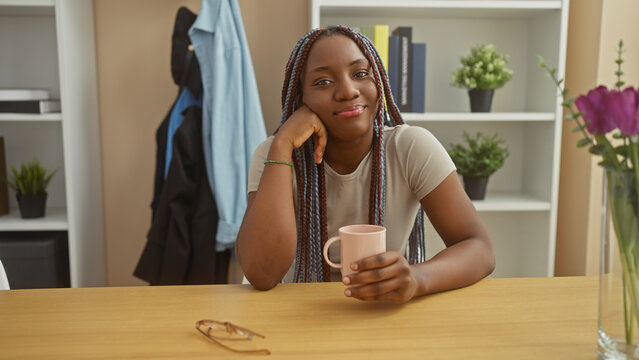 A smiling african-american woman with braids sits at a table in a cozy home interior holding a mug. - Powered by Adobe