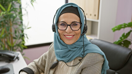 A smiling mature woman wearing a hijab and headset sits in an office, representing professional customer support.