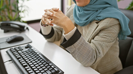 A professional woman with a hijab sitting at an office desk, hands clasped in a modern indoor workplace setting.