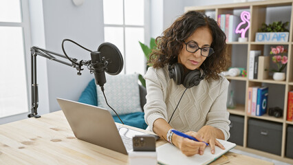 Middle-aged hispanic woman takes notes in radio studio with microphone and laptop.