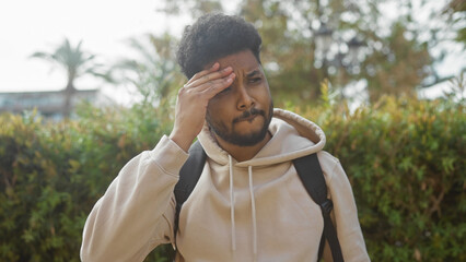 African american man with a beard outdoors wearing a backpack and a hoodie pressing head, stressed or ill, in a park setting.