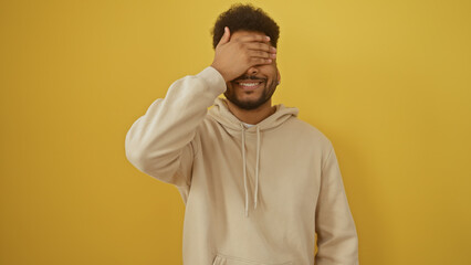 Smiling african american man in casual hoodie covering eyes against yellow background, portraying anticipation or surprise.