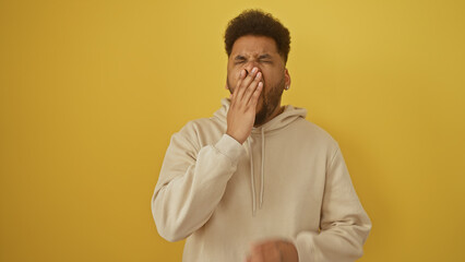 A tired african american man in casual attire yawning against a plain yellow background.