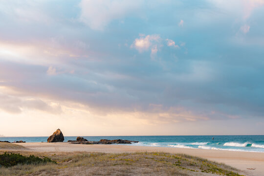 Rocks on Currumbin Beach illuminated at sunset with person windsurf foiling in the distance