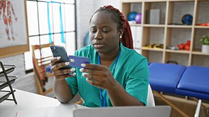 African american woman with braids using smartphone and credit card in rehab clinic interior.
