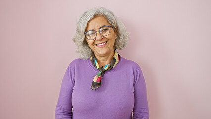 A cheerful middle-aged woman with glasses and grey hair, dressed in purple, stands before a pink background.