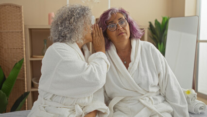 Mature women in white robes whispering in a cozy spa room indicate friendship and relaxation