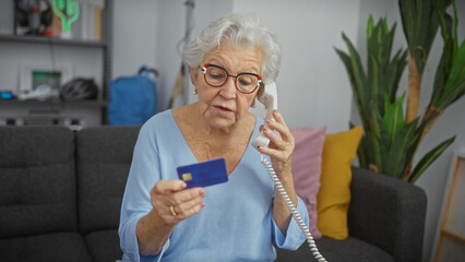 Concerned elderly woman holding credit card and telephone in her apartment living room. - Powered by Adobe