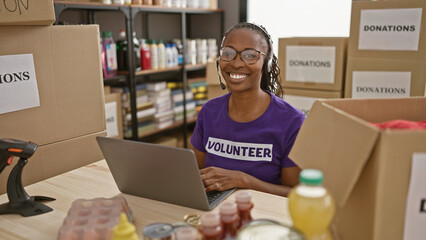 Smiling woman volunteering at a donation center, using a laptop amid boxes and supplies.