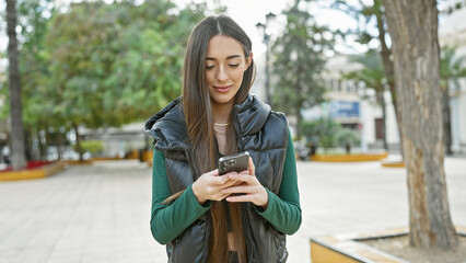 A young woman texting on her smartphone in a city park during daytime