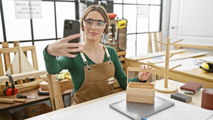Woman carpenter taking selfie with smartphone in sunny woodworking workshop, showcasing handmade wooden organizer.