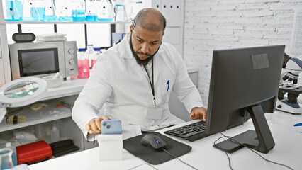 African man working in a laboratory using a computer and a smartphone, indoors.