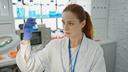 A focused young woman scientist examines a blue liquid in a flask in a well-equipped laboratory.