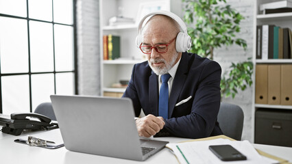 Obraz premium A senior businessman in headphones using a laptop in a modern office, implying telecommuting or online communication.