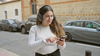 A young hispanic woman enjoys music on her headphones while using her smartphone on a city street.