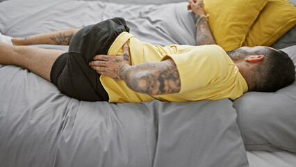 Hispanic man with tattoos lying in bed with back pain in a modern bedroom setting.