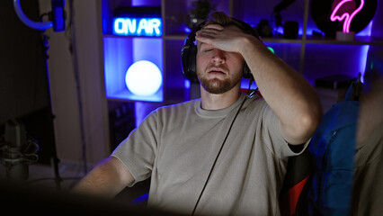 A stressed young man with a beard in a dark gaming room, wearing headphones and touching his forehead, exuding frustration or concentration. © Krakenimages.com