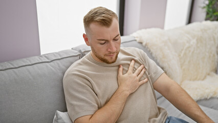 Young caucasian man experiencing chest pain on a couch in a home interior.