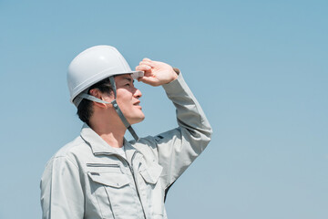青空の下でヘルメットを被って空を見上げる作業着姿のアジア人男性作業員
