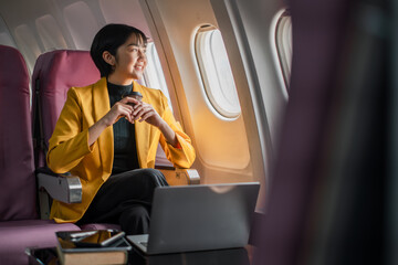 Professional woman in a yellow blazer working on a laptop and looking out the window while traveling on an airplane.