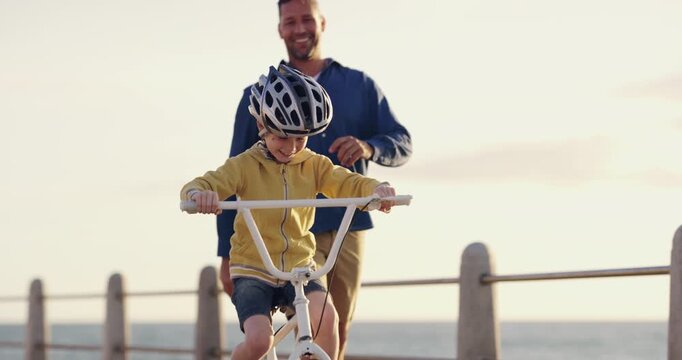 Learning, bicycle and proud dad teaching his young son to ride while wearing a helmet for safety along a seaside promenade. Active father helping and supporting his child while cycling at sunset