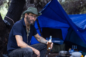 homeless man sitting at his secluded camping spot under trees drinking beer