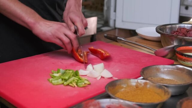 Close-up of a chef's hands chopping tomatoes, bell peppers and onions on a professional cutting board surrounded by cumin and turmeric seasonings for preparing Uzbek meat pilaf.