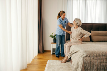 Nurse is helping a senior woman to stand up from a bed during her home visit.