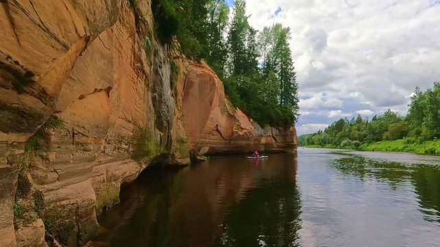 Kayakers paddle along the serene river with rocky cliffs and lush greenery in Gauja National Park, Latvia