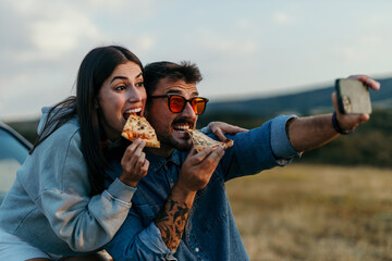 Loving couple taking a selfie outdoors, leaning on the hood of their car in the evening, eating fast food