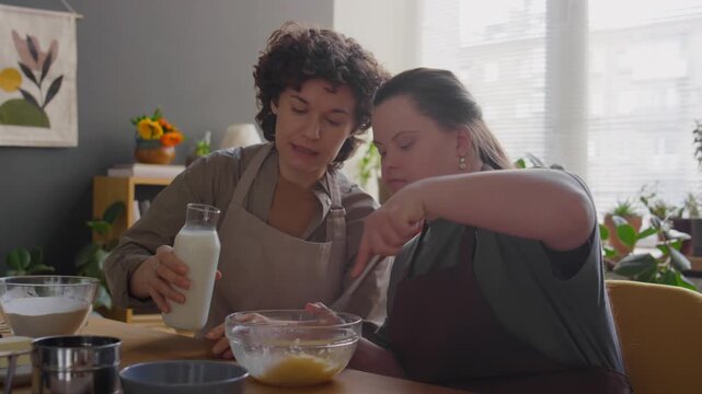 Female teacher adding milk to glass bowl while girl with Down syndrome mixing ingredients for baking on cooking class