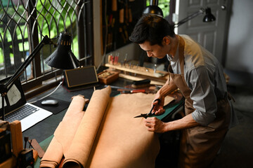 Leather craftsman cutting a large piece of leather with scissors on a workbench. Handicraft business concept