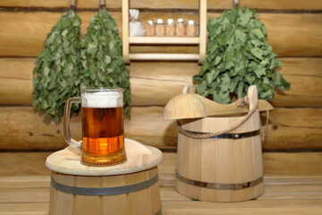 On a wooden bucket in the sauna interior is a glass mug of light beer on the background of a log wall with dry birch and oak brooms and shelf with set of aroma oil bottles.