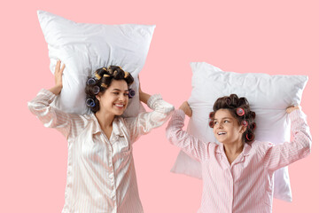 Little girl and her mother in pajamas with hair curlers holding pillows on pink background