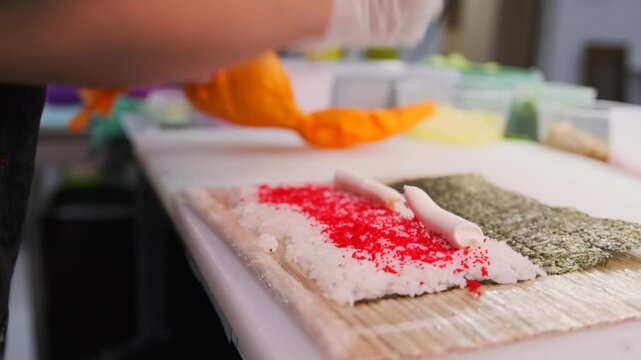 Side view of a chef's hands preparing sushi maki rolls using curd cheese red caviar masago rice and a Nori sheet lying on a bamboo mat. Close up.
