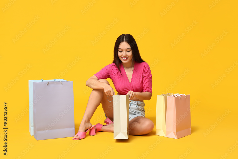 Poster fashionable young asian woman in stylish denim shorts with shopping bags sitting against yellow back