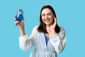 Beautiful young woman holding glass of tasty cocktail with blueberry on blue background