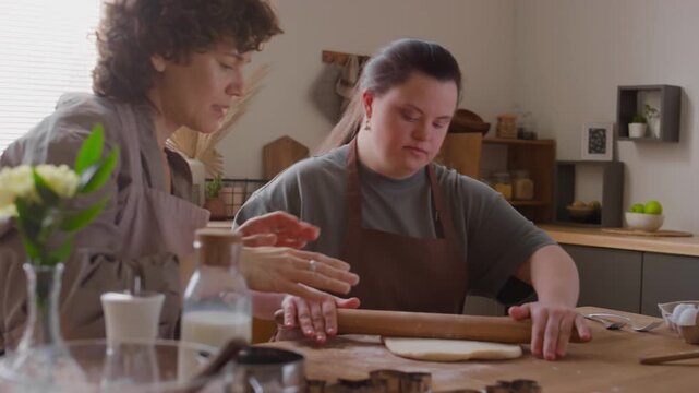 Female tutor teaching girl with Down syndrome to roll dough and sifting flour over rolling pin during process