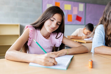 Young caucasian elementary student girl writing in notebook while studying at primary school. Children education concept.