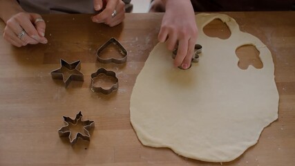 Top down of hands of girl using cookie cutter to cut out pastry dough on kitchen table with help of female teacher