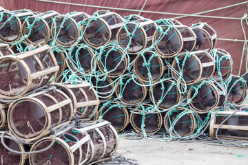 A large stack of cylindrical fishing traps is neatly arranged beside a waterfront dock, ready for use in capturing marine life, with boats and a town in the background.