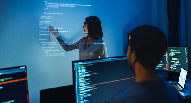 asian woman developers collaborating on a coding project, with one member pointing at code projected on the wall while the other works on multiple computer screens
