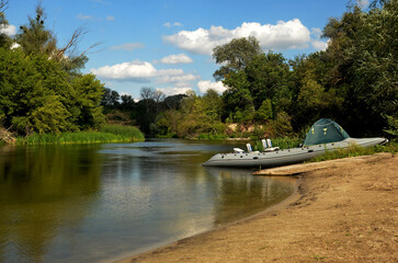 Moored inflatable boat sits peacefully on the river bank while trees form a serene backdrop, creating a piece of environmental art under a blue sky dotted with white clouds.