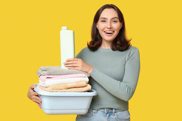 Young woman with laundry basket and bottle of detergent on yellow background