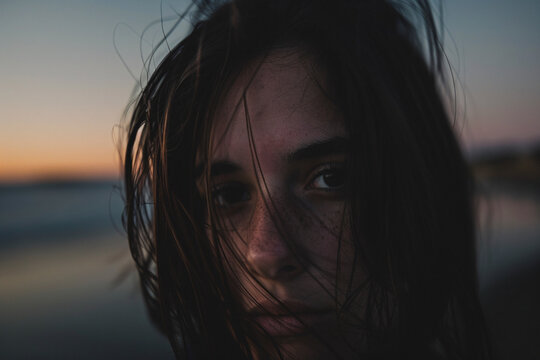 A close-up portrait of a young woman with windblown hair, looking directly at the camera with a sunset backdrop in the background, reflecting a serene and contemplative mood.