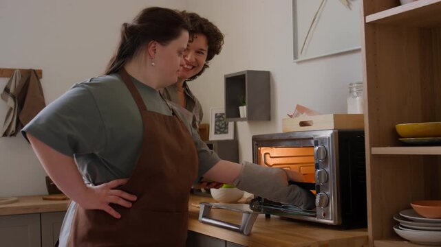 Girl with Down syndrome taking off baking sheet from hot oven while making cookies with female teacher in kitchen