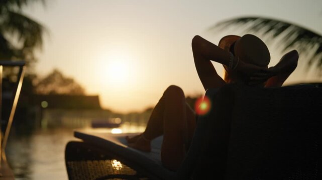 Silhouette of beautiful woman relaxing on deck chair near pool in sunset. Traveling in summer vacation and enjoying life, carefree lady admiring sundown in beautiful tropical garden, tourism in summer