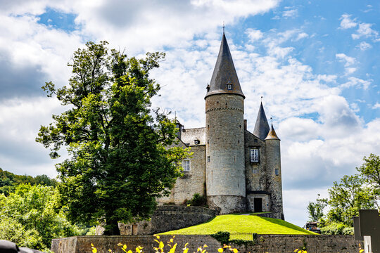 Veves Castle on hill against blue sky, towers with conical roofs and stone walls, a huge tree with green leaves, a sunny day in Houyet, Namur province, Wallonia, Belgium