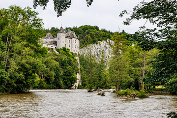 Lesse river and Walzin castle on rocky hill protruding from lush green trees in background, stone walls and towers, stream of water overflowing, sunny summer day in Namur province, Wallonia, Belgium